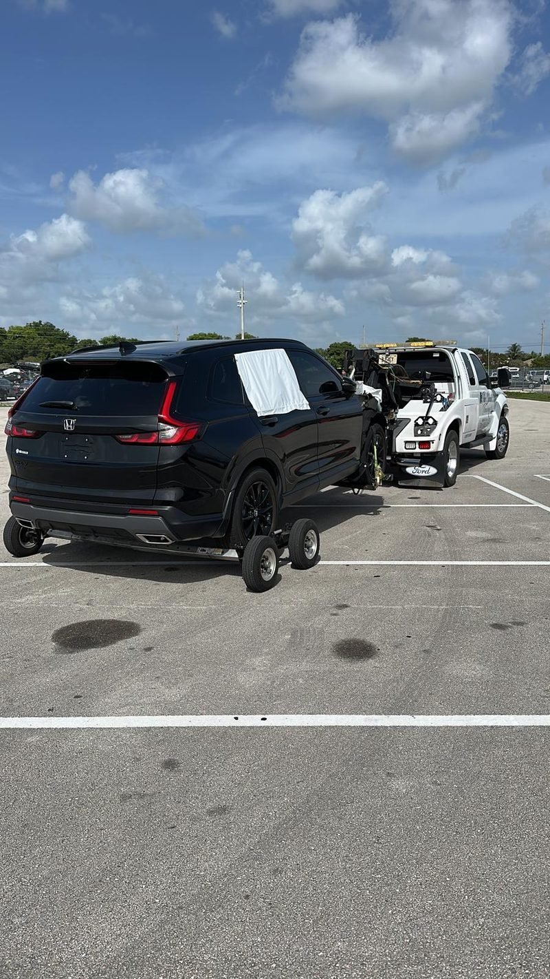 Black Honda CR-V on tow dollies behind the white wrecker in a parking lot under a blue sky