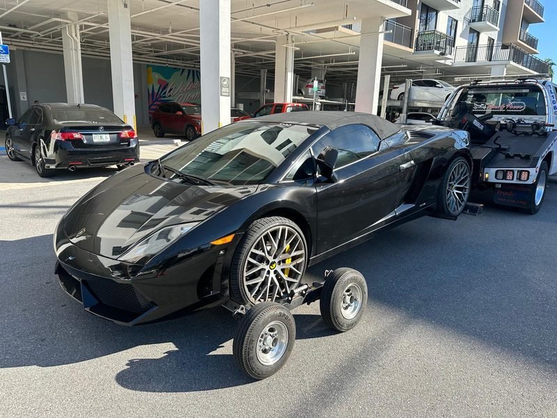 Black Lamborghini Gallardo Spyder resting on tow dollies under a residential parking garage next to the wrecker