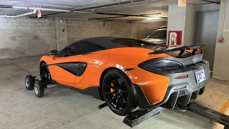 Orange McLaren 600LT on tow dollies inside a low-clearance concrete parking garage